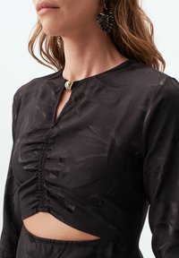 Zwart langslurf shirt met een keyhole halslijn, gerimpelde stof en uitgesneden ontwerp bij de taille. Voorzien van een metallic accent bij de halslijn.