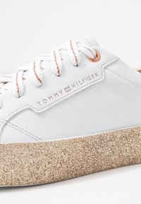 White leather sneaker with pink accents and laces. Features a textured beige rubber sole and "TOMMY HILFIGER" branding on the side.