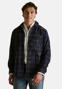 Flanellhemd in marineblau, grün und gelb kariert; verfügt über zwei Brusttaschen, eine knöpfbare Vorderseite und eine weiche Textur. Darunter ist ein weißes Hemd zu sehen.