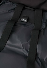 Bolsa de nylon negra con dos correas negras y una etiqueta que dice "MNG." El material tiene una textura lisa y brillante con un ligero brillo.