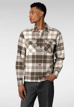 Mannlig modell iført en brun, grå og hvit rutete knappet jakke med brystlommer, står med den ene hånden i lommen og ser til siden.