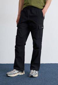 Een persoon staat met één hand in de zak van zwarte cargobroeken, draagt een groen shirt en grijze sneakers op een blauw tapijt.