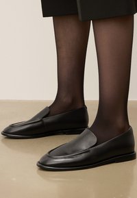 Mocassins en cuir noir au profil élégant, avec des détails cousus et une texture lisse, posés sur des collants noirs transparents.