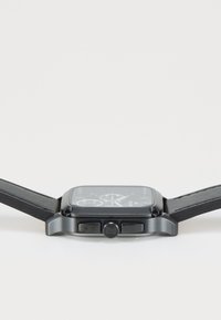 Montre carrée noire avec un boîtier en métal et deux boutons-poussoirs. Elle dispose d'un bracelet en cuir noir et d'un cadran foncé avec des accents argentés.