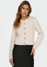 Cardigan colore crema realizzato in tessuto a maglia, con scollatura rotonda, sei bottoni in legno e due tasche frontali. Maniche lunghe, design corto.