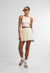 Ljust rosa cropped tank top i kombination med en krämfärgad plisserad kjol och bruna sneakers med rosa accenter. Klara, rektangulära solglasögon bärs.