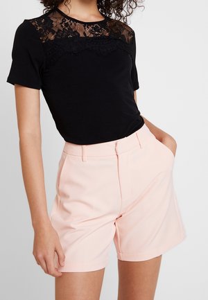 Femme portant un haut noir à manches courtes avec un détail en dentelle et un short taille haute rose clair, une main dans la poche, sur un fond uni.