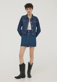 Giubbotto di jeans scuro con due tasche frontali, abbinato a una gonna di jeans scura. Indossato con stivali da cowboy neri, con punta appuntita e tacco basso.