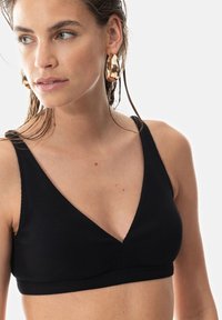 Musta ribbattu bralette, jossa on V-aukko ja leveät hihnat. Kangas on teksturoitunut ja istuva, luoden saumatonta ilmettä. Kultaiset renkaankorvakorut.