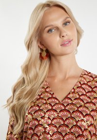 Bluză cu decolteu în V, decorată cu un model de fani roșii și aurii, confecționată dintr-un material moale și ușor. Cercei aurii cu accente texturate și franjuri.