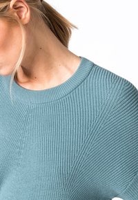 Femme aux cheveux clairs portant un pull en maille côtelée bleu, tête inclinée vers le bas et partiellement tournée de côté sur un fond blanc.