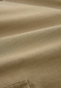 Beige stof med et subtilt ternet mønster, der har en glat tekstur og en folde-sektion, der viser syede detaljer.