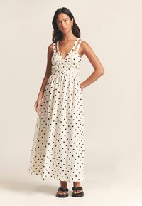 Maxiklänning i krämfärg med svarta prickar, med figurnära överdel, rynkdetaljer och breda axelband; kombinerad med svarta sandaler.