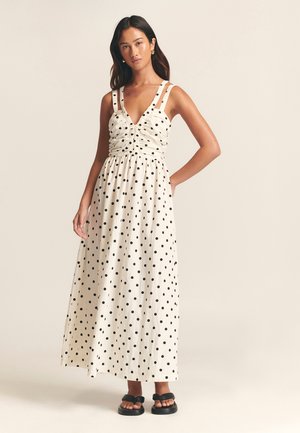 Weißes Maxikleid mit schwarzen Punkten. Hat einen tiefen V-Ausschnitt, ein gerafftes Oberteil, breite Träger und einen schwingenden Rock. Wird mit schwarzen Sandalen getragen.