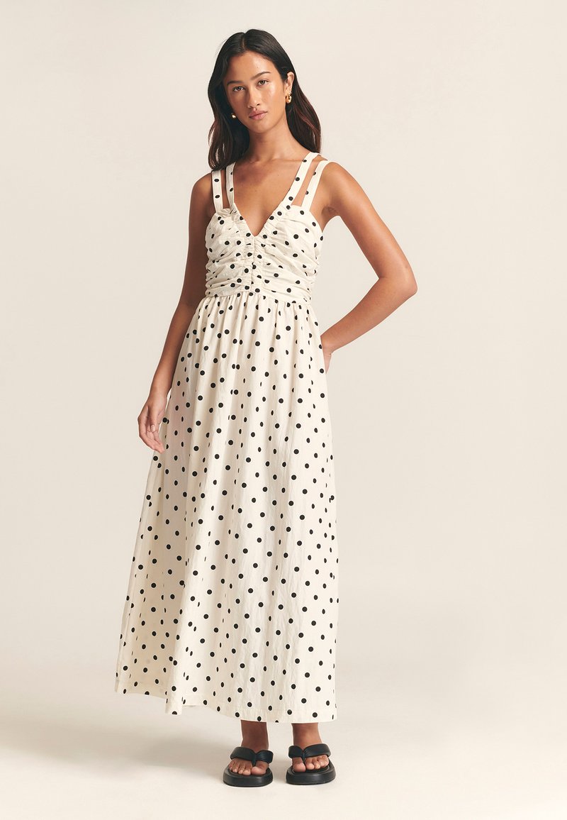 Maxiklänning i krämfärg med svarta prickar, med figurnära överdel, rynkdetaljer och breda axelband; kombinerad med svarta sandaler.