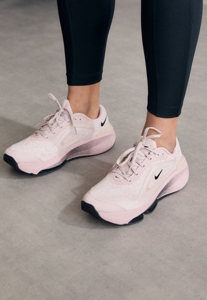Stopy w jasno różowych butach do biegania Nike z czarnymi logo Swoosh, w połączeniu z czarnymi legginsami sportowymi, stojące na szarej podłodze.