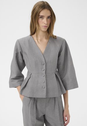 Femme portant une veste grise cintrée avec boutons, manches trois-quarts, et un pantalon plissé assorti, sur un fond uni.