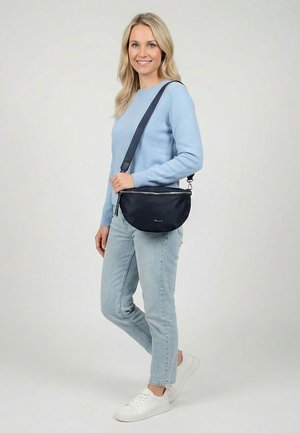 Sieviete gaiši zilā džemperī un džinsos ar melnu pār plecu krustu siksnu, stāv un smaida pret vienkāršu fona virsmu.