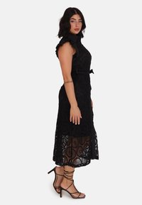 Abito in pizzo nero con motivo circolare ricamato, corpetto aderente, maniche arricciate, vita sfilata e parte inferiore semi-trasparente. Décolleté con cinturini.