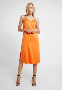 Žena v oranžových midi šatách bez rukávov s bočnými rozparkami a čiernymi sandálmi na vysokom opätku s remienkom okolo členka, stojaca pred jednoduchým bielym pozadím.