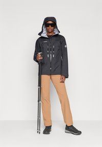 Giacca impermeabile nera con cappuccio, frontale con zip e tasche, indossata con pantaloni beige e robuste scarpe da trekking nere. Tiene dei bastoni da trekking.