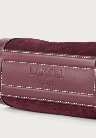 Крупен план на бордо велурена и кожена чанта с бели шевове и релефно лого Lancel Paris.