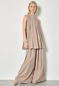Mujer con túnica sin mangas de tono topo suelta y plisada y pantalones de pierna ancha, con tacones beige y pulseras crema, de pie contra una pared lisa.
