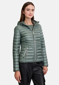 Groene pufferjack met horizontaal gestikte ontwerp, aangesloten snit, ritszakken en verstelbare capuchon. Draagt over een donkere top en broek.