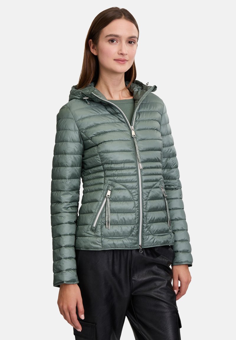 Groene pufferjack met horizontaal gestikte ontwerp, aangesloten snit, ritszakken en verstelbare capuchon. Draagt over een donkere top en broek.