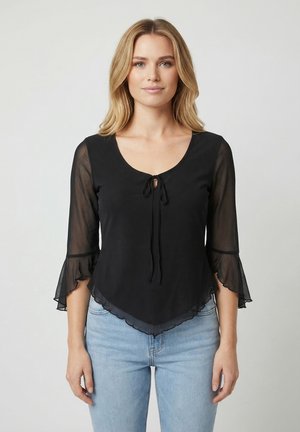 Donna con capelli biondi che indossa una maglia nera trasparente con maniche e fiocco davanti e jeans azzurri, in piedi contro uno sfondo chiaro uniforme.