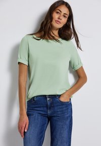 Haut vert clair à manches courtes avec des poignets roulés, fait d'un tissu doux. Associé à un jean en denim bleu, affichant une coupe décontractée.
