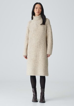 Cremfarbendes, gestricktes Sweatkleid mit hohem Kragen, langen Ärmeln und geripptem Saum; getragen über schwarzen Strumpfhosen und braunen Stiefeletten.