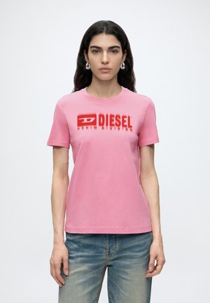 Frau mit langen dunklen Haaren, die silberne Creolen, ein pinkes Diesel-T-Shirt und blaue Jeans trägt, steht vor einem schlichten Hintergrund.