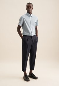 Korte mouwen, lichtblauw geruit shirt met knoopkraag, gecombineerd met donkere geplooide broek en zwarte leren schoenen. Neutrale achtergrond.