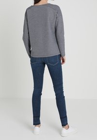 Felpa grigia con cuciture delle spalle scese, abbinata a jeans skinny blu scuro. Il modello indossa sneaker bianche. Sfondo neutro.