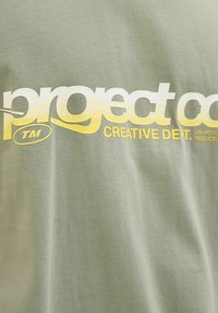 Maslinasto zelena pamučna majica s velikim, teksturiranim, bijelim i žutim grafičkim natpisom koji glasi "project co. CREATIVE DEPT."
