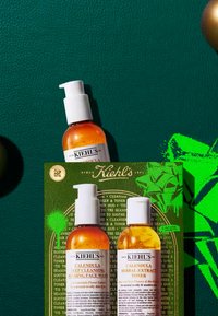 Zestaw kosmetyków Kiehl's z dwoma bursztynowymi butelkami z białymi pompkami, umieszczonymi na ciemnozielonym tle. Z przodu karta z grafiką zielonych roślin.