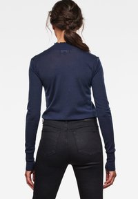 Suéter de manga larga azul marino con cuello redondo y puños acanalados, combinado con jeans negros ajustados que presentan detalles de costura en los bolsillos traseros.