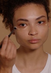 Application de maquillage avec un petit pinceau sur la zone sous les yeux, présentant des points de correcteur de teint, sur un fond neutre.