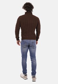 Maglione lavorato a maglia marrone con un alto colletto, texture a coste e maniche lunghe, abbinato a jeans azzurri chiari e sneaker bianche.