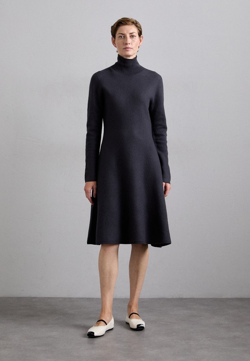 Dunkelgraues, langärmeliges Kleid mit hohem Kragen, A-Linien-Silhouette und weicher Textur; kombiniert mit weißen Schuhen mit schwarzem Zehenkappen.