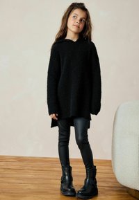 Haut à capuche oversized noir en tissu texturé, associé à des leggings en cuir noir et des bottines avec boucles. Fond en parquet en bois.