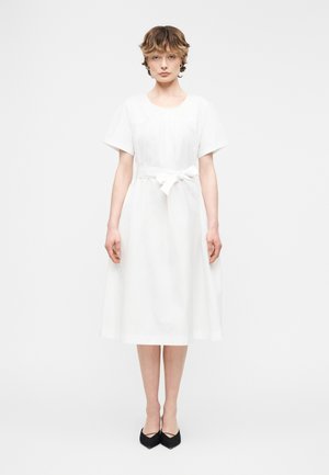 Kobieta stojąca przodem, ubrana w białą midi sukienkę z krótkim rękawem i paskiem w talii oraz czarne szpilki z zaostrzonym noskiem na białym tle.
