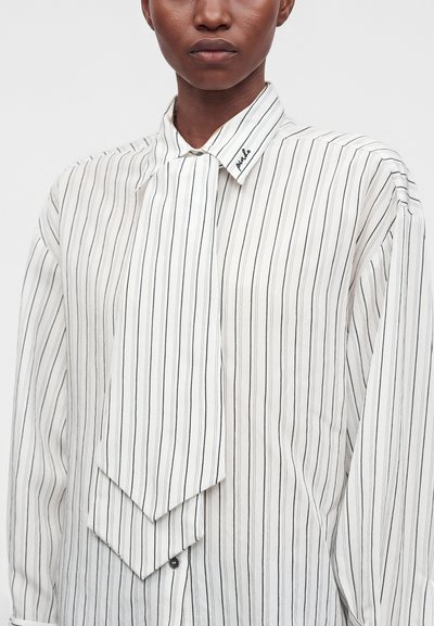 Personne portant une chemise blanche à boutons avec de fines rayures noires verticales et une large cravate du même motif.