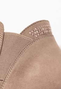 Detailní záběr na béžovou semišovou patu boty s elastickým bočním panelem a malými růžovými rhinestonovými ozdobami blízko horního okraje.