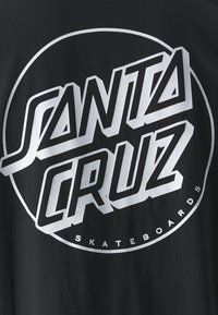Juodas marškinėliai su dideliu, ryškiu baltu grafiką, kuriame užrašyta "SANTA CRUZ" ir "SKATEBOARDS" stilizuotu šriftu, įkomponuota apskritime.