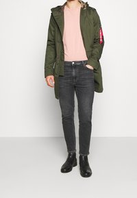 Parka verde con forro de camuflaje, camiseta de color rosa pálido, jeans ajustados negros y botas de tobillo negras. Tiene bolsillos y un detalle de insignia en la manga.