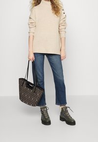Beżowy sweter dziany z wysokim kołnierzem, niebieskie przylegające dżinsy, ciemnozielone buty za kostkę sznurowane, oraz brązowa torba z wzorem z czarnymi uchwytami.