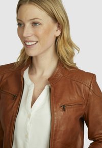 Chaqueta de cuero marrón con un diseño ajustado, presenta un cuello alzado, detalles acolchados en los hombros y bolsillos con cremallera. Llevada sobre una camisa blanca.