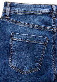 Denim-farkut tummansinisinä, joissa on kulmikas takatasku ja kontrastivärinen oranssi ommel saumoissa ja reunoissa.